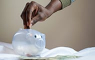 a person of color with a green sleeve inserts a coin into a clear piggy bank